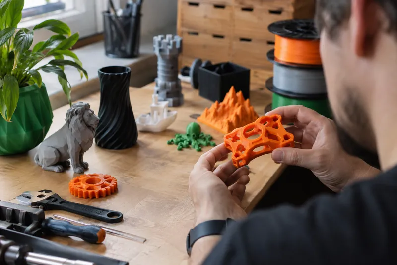 Oranžový díl z Original Prusa MK4S v rukou tvůrce mezi 3D výtisky a filamenty